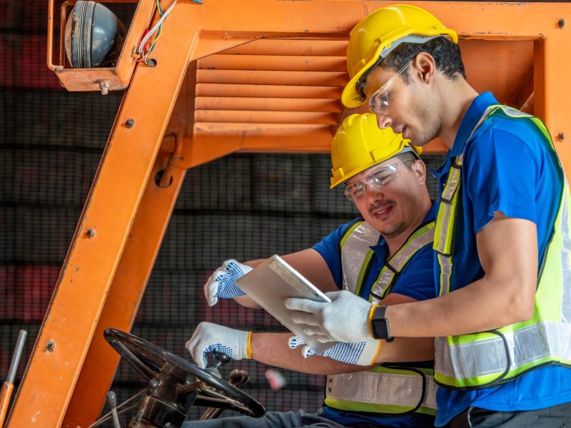 Two warehouse workers wearing hard hats and safety vests are using a tablet computer to check inventory while sitting on a forklift.